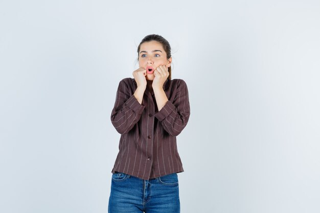 Young girl with fists near mouth, keeping mouth open in striped shirt, jeans and looking shocked. front view.