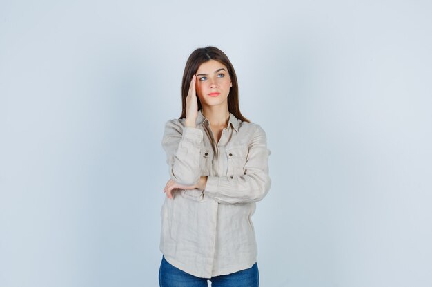 Young girl with hand on temple, thinking about something in beige shirt, jeans and looking pensive. front view.