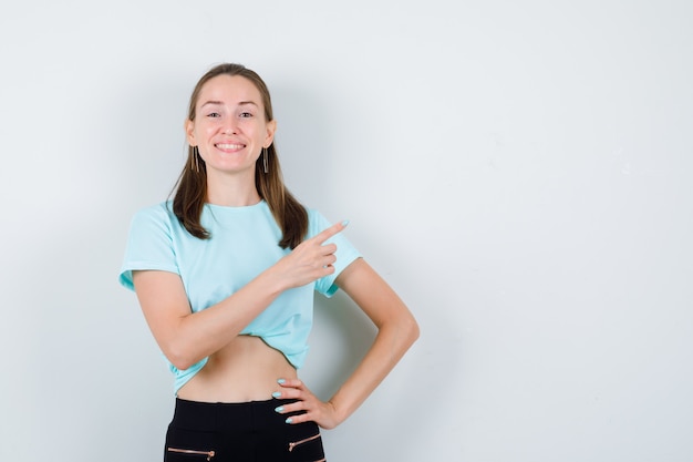 Young girl with hand on waist, pointing up in turquoise t-shirt, pants and looking cheerful , front view.