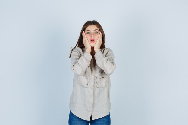 Young girl with hands on cheeks, keeping mouth open in beige shirt, jeans and looking surprised , front view.