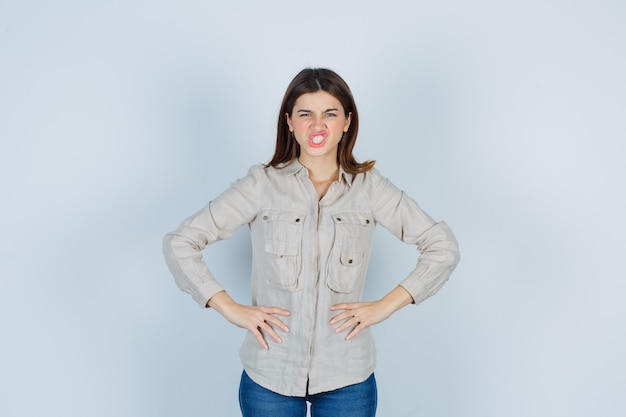 Free photo young girl with hands on hips in beige shirt, jeans and looking agitated. front view.