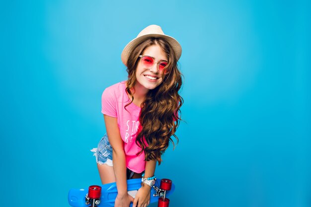 Young girl with long curly hair in pink sunglasses posing on blue background in studio. She wears shorts, pink T-shirt, hat,. She holds blue skateboard  and smiles to camera.