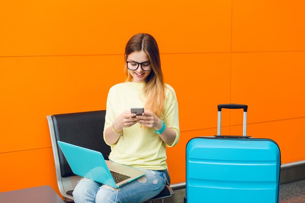 Young girl with long hair and black glasses is sitting on chair on orange background. She wears jellow sweater with jeans. She has laptop on knees and suitcase near. She is typing on phone.