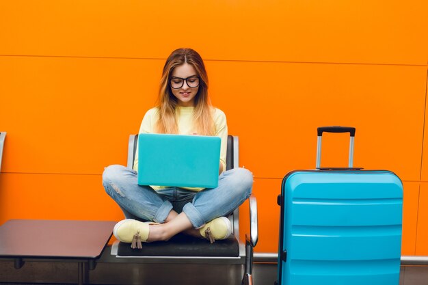 Young girl with long hair is sitting on chair on orange background. She wears yellow sweater and jeans. She is typing on laptop.