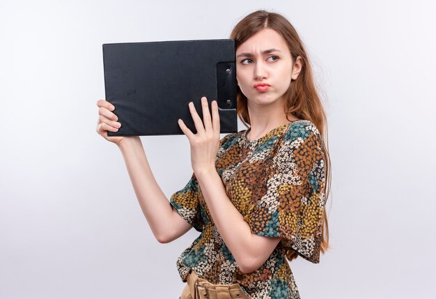 Young girl with long hair wearing colorful dress holding clipboard looking aside with frowning face 