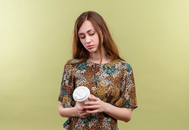 Молодая девушка с длинными волосами в ярком платье держит чашку кофе, глядя на нее с грустным выражением лица
