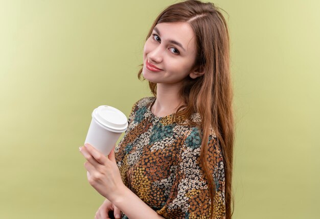 Молодая девушка с длинными волосами в ярком платье держит чашку кофе, уверенно улыбаясь