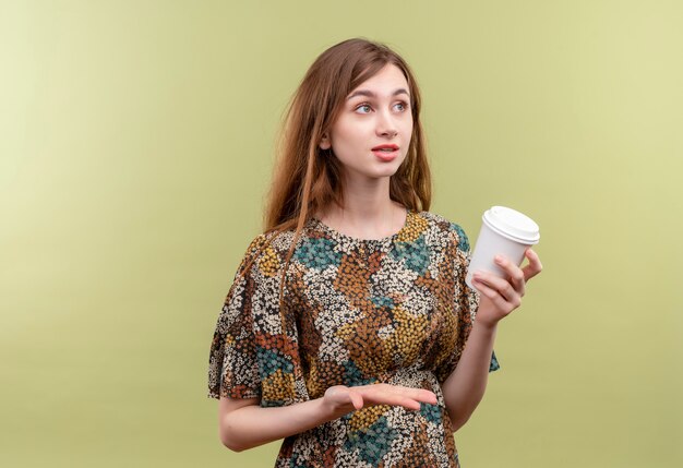 Young girl with long hair wearing colorful dress holding coffee cup smiling looking aside confused  