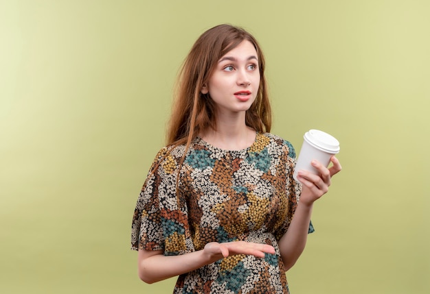 Молодая девушка с длинными волосами, одетая в красочное платье, держит чашку кофе, улыбаясь в замешательстве