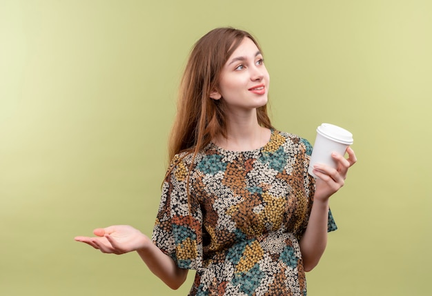 Молодая девушка с длинными волосами, одетая в красочное платье, держит чашку кофе, улыбаясь, глядя в сторону, разводя руку в сторону
