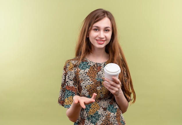 Молодая девушка с длинными волосами, одетая в красочное платье, держит чашку кофе, улыбаясь, глядя на камеру, поднимая руку, задавая вопрос
