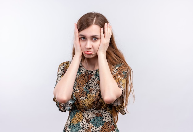 Young girl with long hair wearing colorful dress looking diappointed with unhappy face touching with arms her face 