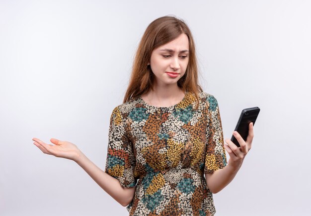 Молодая девушка с длинными волосами в ярком платье смотрит на экран мобильного телефона с растерянным выражением лица, разводя руку в сторону