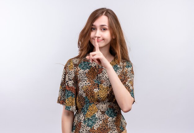 Young girl with long hair wearing colorful dress making silence gesture with finger on lips 