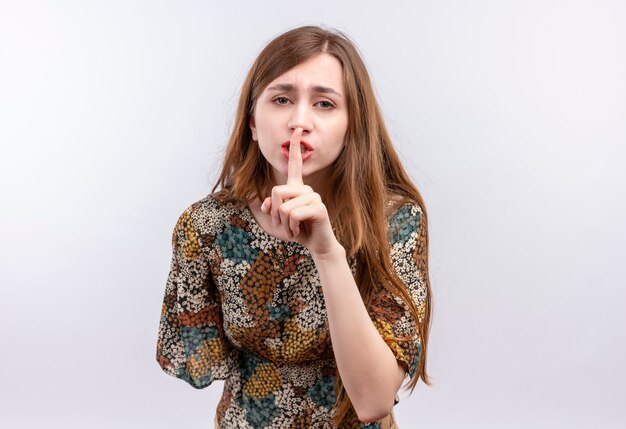 Young girl with long hair wearing colorful dress making silence gesture with finger on lips 