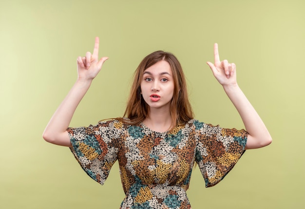 Giovane ragazza con i capelli lunghi che indossa abiti colorati che punta il dito indice verso l'alto con seria espressione fiduciosa sul viso