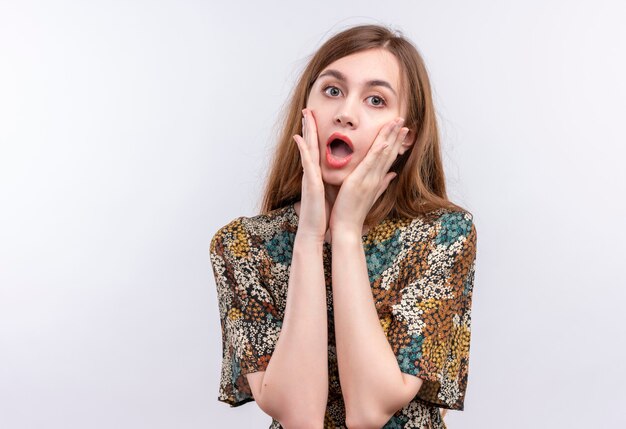 Young girl with long hair wearing colorful dress shocked touching face with arms 