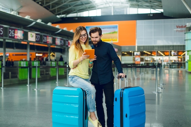 Young girl with long hair in yellow sweater, jeans is sitting on suitcase in airport. Guy with beard in black shirt with pants and suitcase is standing near. They are looking on tablet.