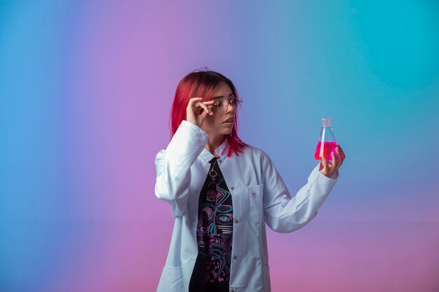 Молодая девушка с розовыми волосами держит химическую колбу и внимательно смотрит.