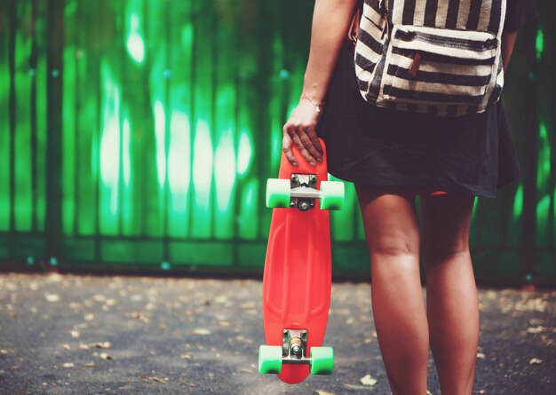 Young girl with plastic orange penny shortboard behind green wall in cap