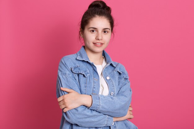 Young girl with pleasant appearance standing against pink wall, dresses denim jacket and white shirt