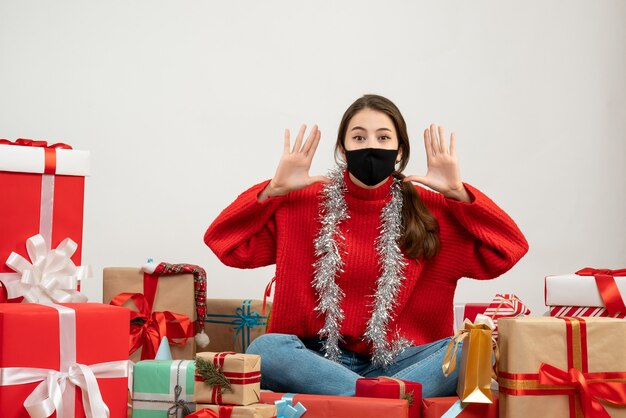赤いセーターと黒いマスクの若い女の子が白いプレゼントの周りに座っている誰かを呼び出します