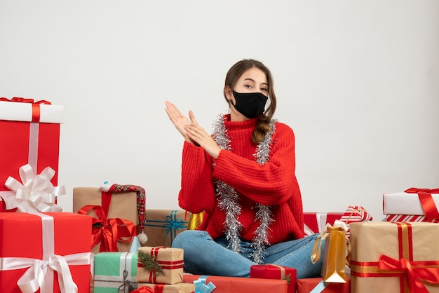молодая девушка в красном свитере и черной маске хлопает в ладоши, сидя вокруг подарков на белом
