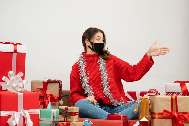 young girl with red sweater and black mask giving hand sitting around presents on white