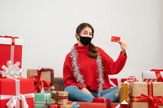 молодая девушка в красном свитере и черной маске с кредитной картой сидит с подарками на белом