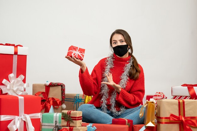 白いセーターとプレゼントの周りに座って贈り物を保持している黒いマスクを持つ少女