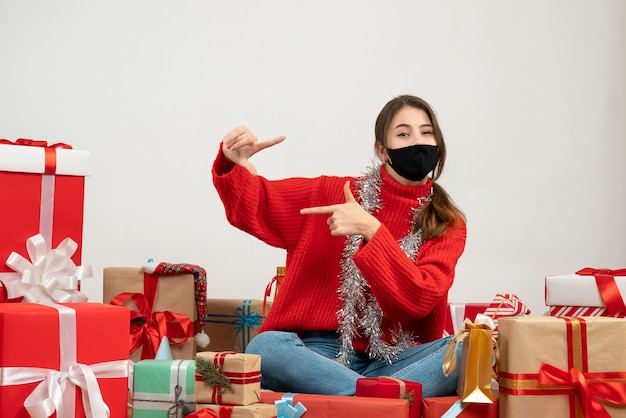 молодая девушка в красном свитере и черной маске делает знак камеры, сидя с подарками на белом