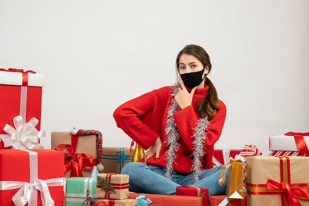 赤いセーターと黒いマスクを持つ少女は、白いプレゼントの周りに座って指銃のサインを作る