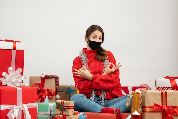 빨간 스웨터와 흰색 선물 주위에 앉아 좋아요 기호를 만드는 검은 마스크 어린 소녀