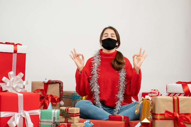 young girl with red sweater and black mask making okey sign sitting around presents on white