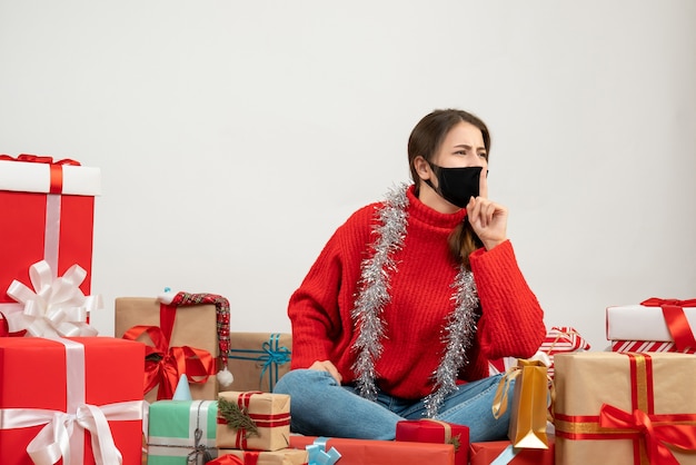 young girl with red sweater and black mask making shh sign sitting around presents on white