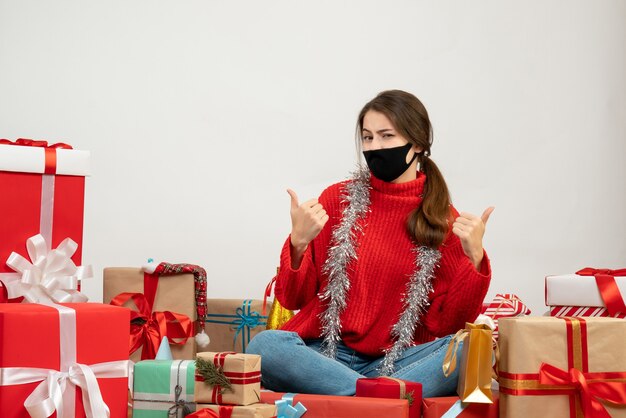 빨간 스웨터와 검은 마스크 엄지 손가락 주위에 앉아 기호를 만드는 어린 소녀 흰색 선물
