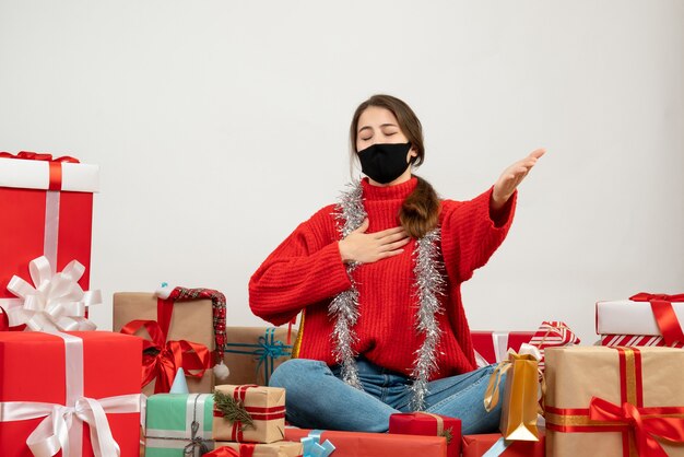 young girl with red sweater and black mask opening hand closing eyes sitting around presents on white