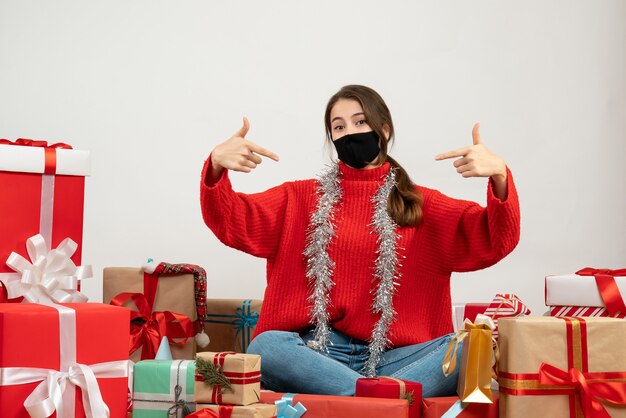 young girl with red sweater and black mask pointing at herself sitting around presents on white
