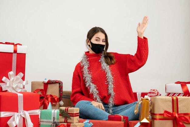 молодая девушка в красном свитере и черной маске, подняв руку, сидит с подарками на белом