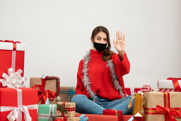 young girl with red sweater and black mask raising her hand sitting around presents on white