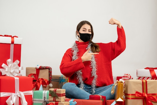 赤いセーターと黒いマスクを持つ少女は、白いプレゼントの周りに座っている彼女の筋肉を披露