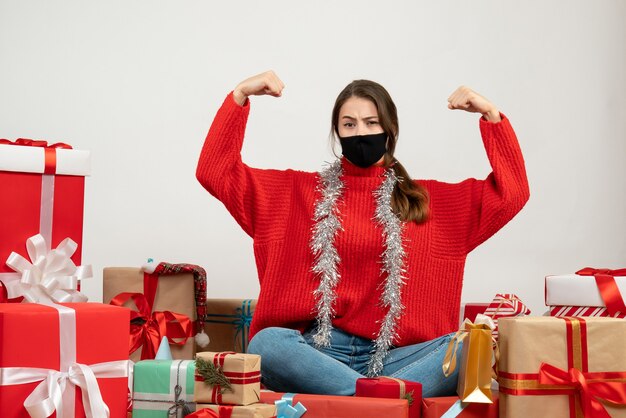 young girl with red sweater and black mask showing off muscles sitting around presents on white