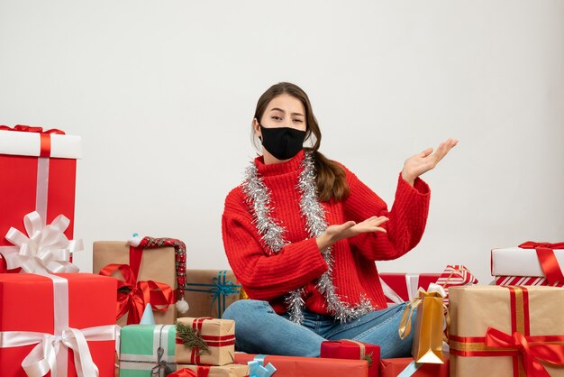young girl with red sweater and black mask showing something sitting around presents on white