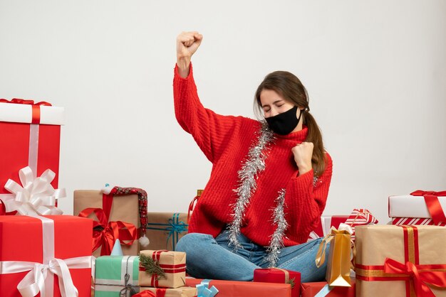 young girl with red sweater and black mask showing winning gesture sitting around presents on white