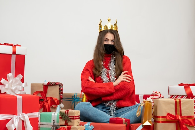 young girl with red sweater crossing hands sitting around presents with black mask on white