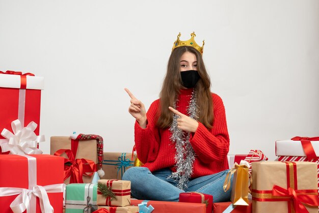 young girl with red sweater finger pointing something sitting around presents with black mask on white