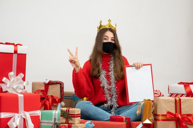 молодая девушка в красном свитере держит файлы, делая V-жест, сидя вокруг подарков с черной маской на белом