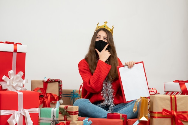 young girl with red sweater holding files putting hands on her chin sitting around presents with black mask on white