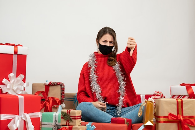 Foto gratuita giovane ragazza con maglione rosso che fa il pollice verso il basso segno seduto intorno presenta con maschera nera su bianco