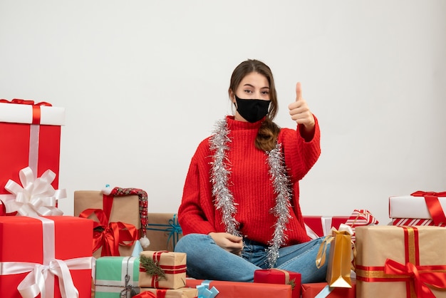 young girl with red sweater making thumb up sign sitting around presents with black mask on white